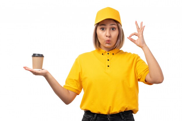 A front view female courier in yellow shirt yellow cap and black jeans holding coffee cup on white