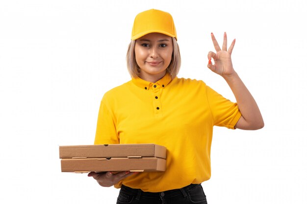 Вид спереди курьер женского пола в желтой рубашке желтой кепке черные джинсы, держа коробки для пиццы, улыбаясь на белом