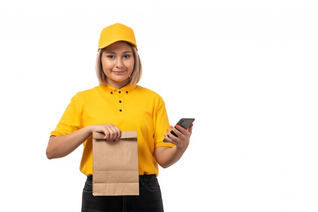 Вид спереди курьер женского пола в желтой рубашке желтой кепке и черных джинсах держит смартфон и пакет с едой на белом