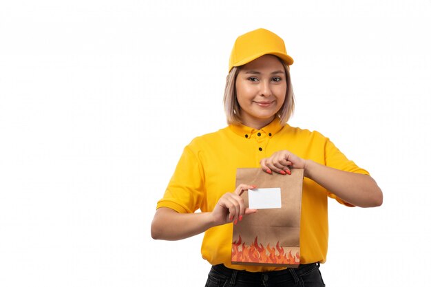A front view female courier in yellow shirt yellow cap black jeans holding white card package smiling on white