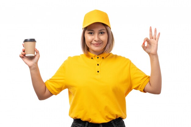 A front view female courier in yellow shirt yellow cap and black jeans posing holding coffee and smiling on white