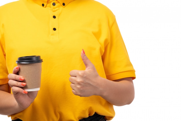 A front view female courier in yellow shirt yellow cap and black jeans posing holding coffee on white