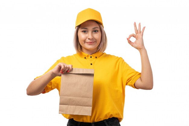 A front view female courier in yellow shirt yellow cap and black jeans posing holding food package smiling on white