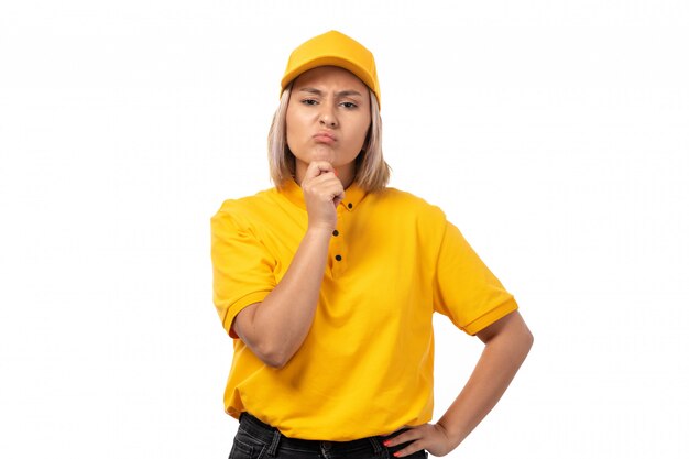 A front view female courier in yellow shirt yellow cap and black jeans posing thinking on white