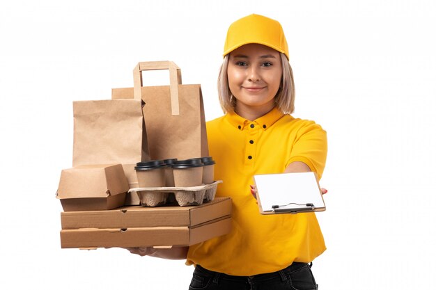 A front view female courier in yellow shirt yellow cap black jeans smiling holding boxes and coffee with paper on white