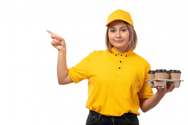 A front view female courier in yellow shirt yellow cap and black jeans smiling holding coffee cups on white