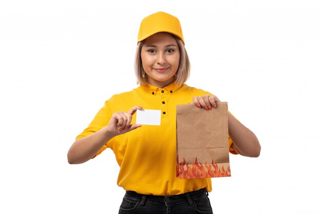 Вид спереди курьер женского пола в желтой рубашке желтой кепке черные джинсы, улыбаясь держа белую карточку и пакеты с едой на белом