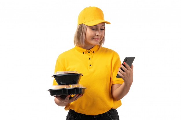 A front view female courier in yellow shirt and yellow cap holding bowls with food and smarpthone smiling on white