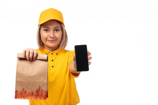 A front view female courier in yellow shirt yellow cap holding smartphone and package with food smiling on white