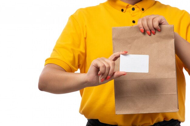 A front view female courier in yellow shirt yellow cap holding white card and package with food on white