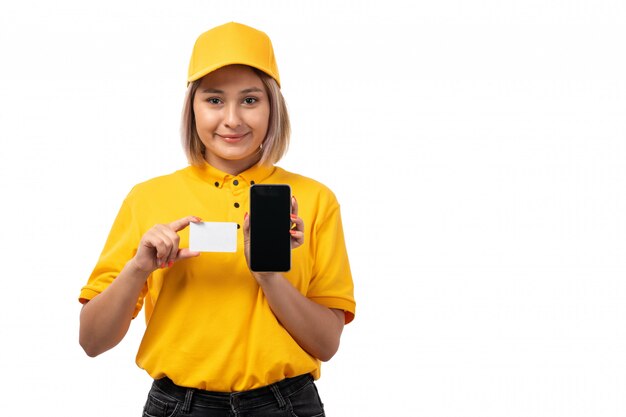A front view female courier in yellow shirt and yellow cap holding white card and smartphone on white