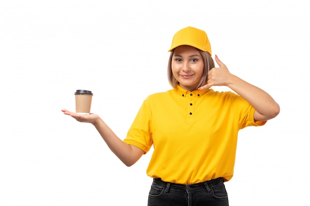 A front view female courier in yellow shirt yellwo cap and black jeans smiling posing holding coffee cup on white uniform