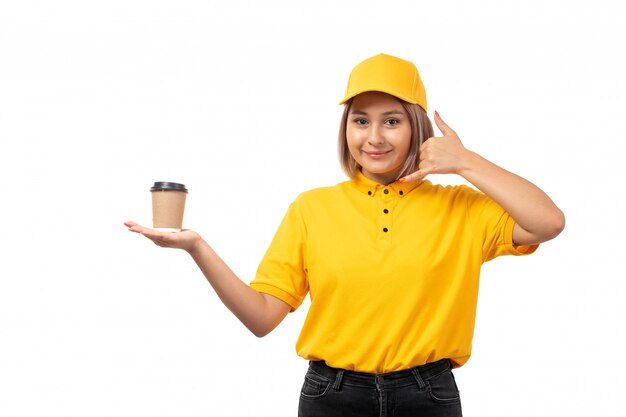 A front view female courier in yellow shirt yellwo cap and black jeans smiling posing holding coffee cup on white uniform
