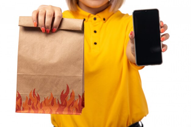 A front view female courier in yellow shrit holding smartphone and food package on white uniform