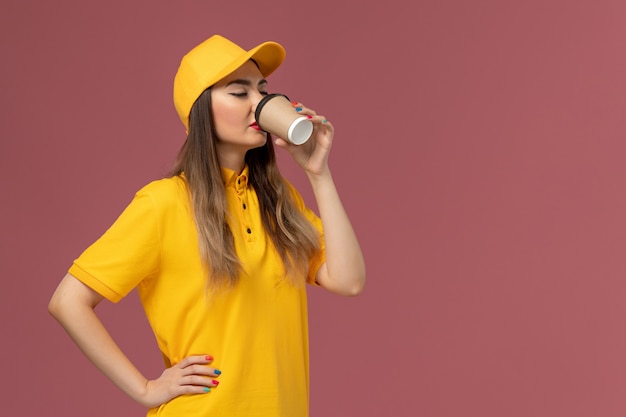 Vista frontale del corriere femminile in uniforme gialla e berretto che beve caffè sulla parete rosa