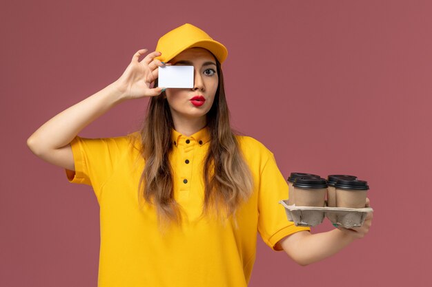 Front view of female courier in yellow uniform and cap holding brown coffee cups and card on the pink wall