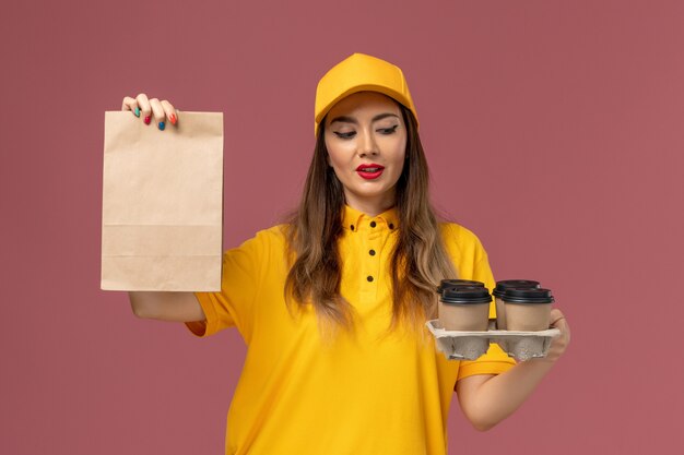 분홍색 벽에 갈색 커피 컵과 음식 패키지를 들고 노란색 유니폼과 모자 여성 택배의 전면보기