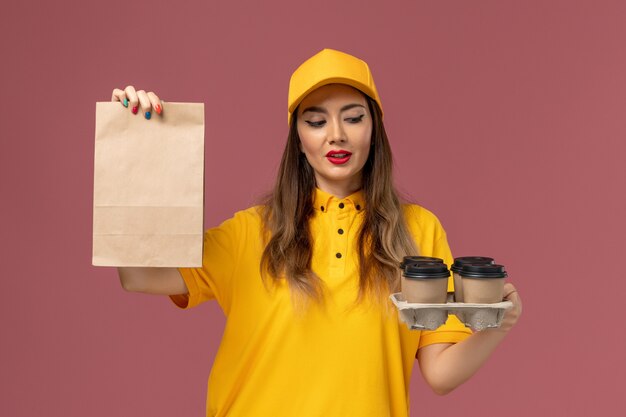 ピンクの壁に茶色のコーヒーカップと食品パッケージを保持している黄色の制服と帽子の女性の宅配便の正面図