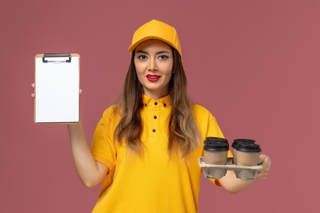 Front view of female courier in yellow uniform and cap holding brown coffee cups and notepad on the pink wall