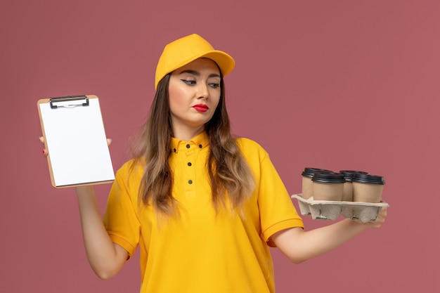 Front view of female courier in yellow uniform and cap holding brown coffee cups and notepad on the pink wall