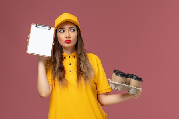 Front view of female courier in yellow uniform and cap holding brown coffee cups and notepad thinking on the pink wall