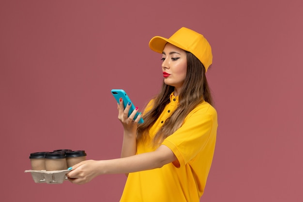 Front view of female courier in yellow uniform and cap holding brown coffee cups and taking photo on pink wall