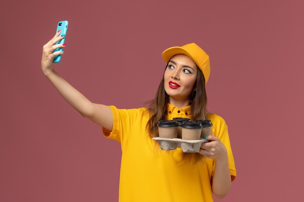 Front view of female courier in yellow uniform and cap holding brown coffee cups and taking a selfie on the pink wall