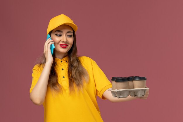 Front view of female courier in yellow uniform and cap holding brown coffee cups and talking on the phone on pink wall