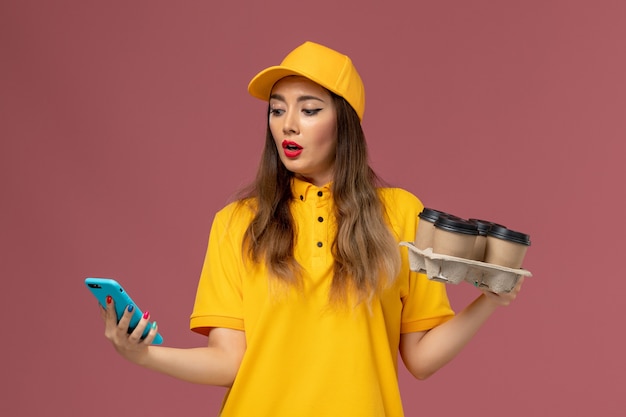 Front view of female courier in yellow uniform and cap holding brown coffee cups and using her phone on the pink wall