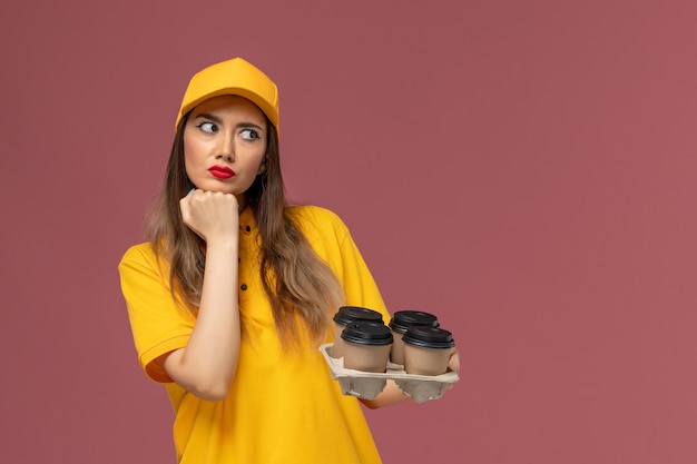 Front view of female courier in yellow uniform and cap holding brown delivery coffee cups and deeply thinking on the pink wall