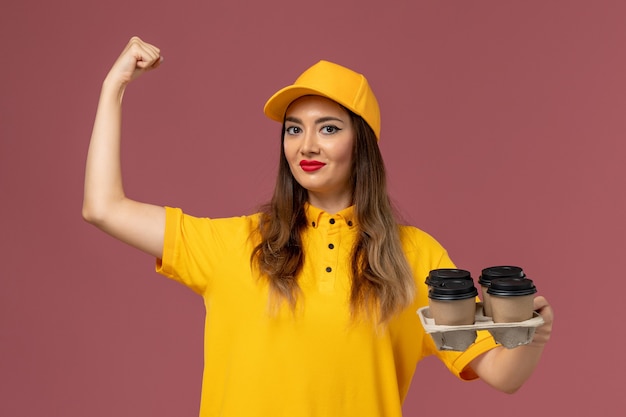 분홍색 벽에 구부러진 갈색 배달 커피 컵을 들고 노란색 유니폼과 모자 여성 택배의 전면보기