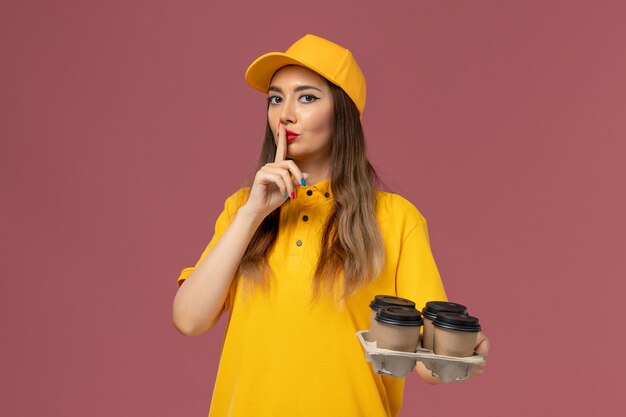 Front view of female courier in yellow uniform and cap holding brown delivery coffee cups on pink wall