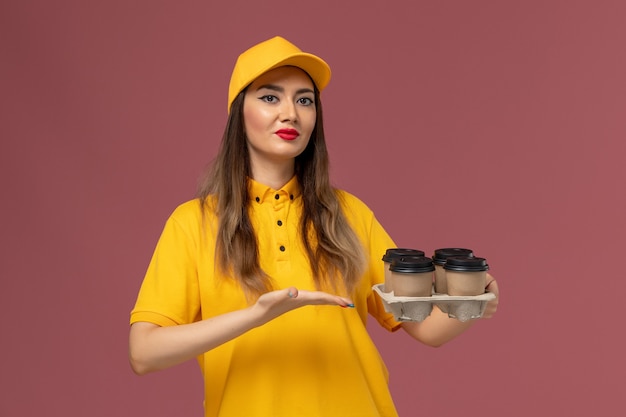 Front view of female courier in yellow uniform and cap holding brown delivery coffee cups on pink wall