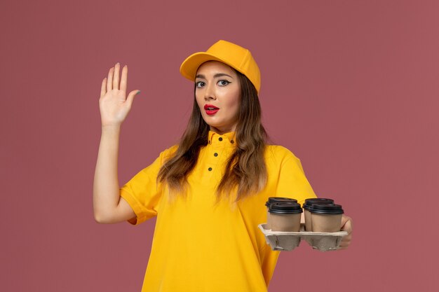 Front view of female courier in yellow uniform and cap holding brown delivery coffee cups on pink wall