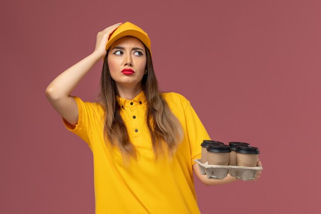 Вид спереди курьера-женщины в желтой форме и кепке, держащего коричневые кофейные чашки на розовой стене