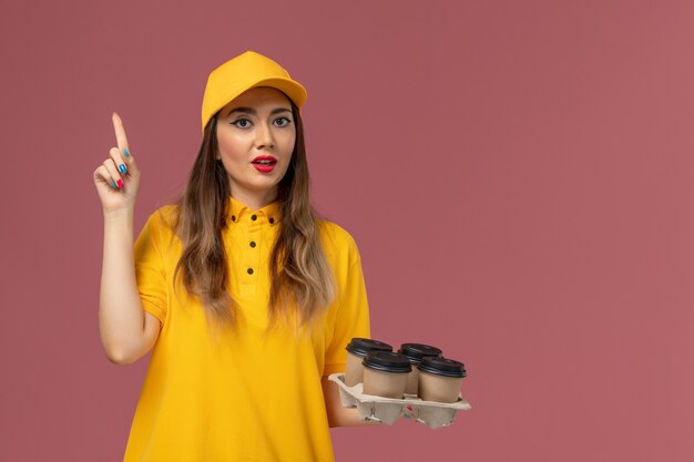 Front view of female courier in yellow uniform and cap holding brown delivery coffee cups on the pink wall