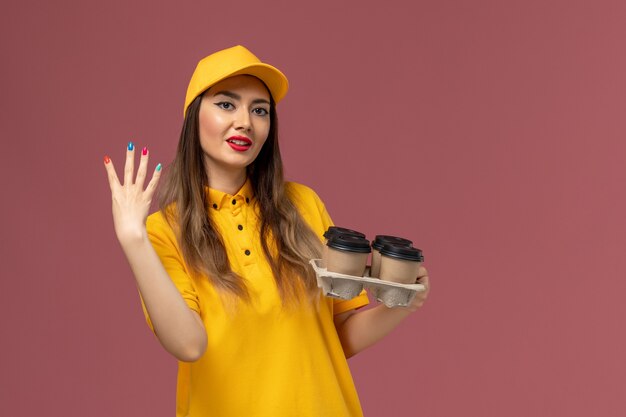 Front view of female courier in yellow uniform and cap holding brown delivery coffee cups on the pink wall