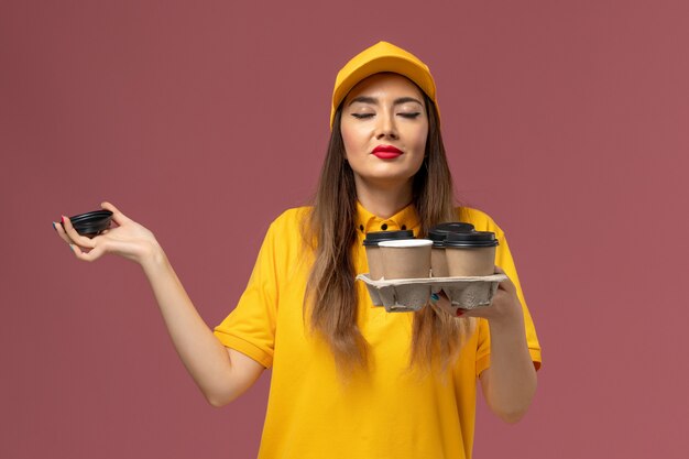 Free photo front view of female courier in yellow uniform and cap holding brown delivery coffee cups smelling scent on pink wall