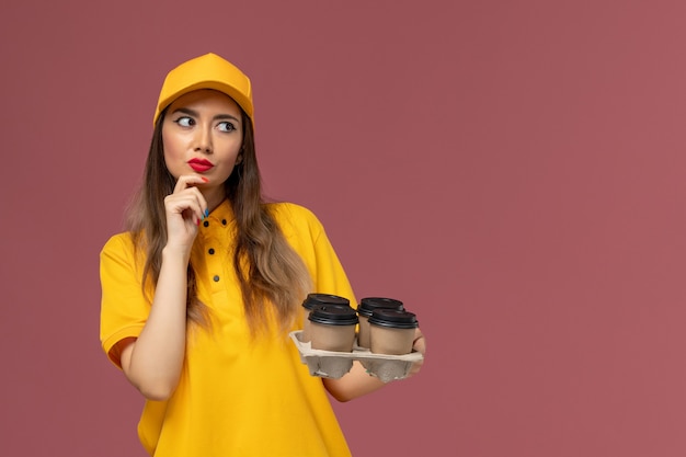 Front view of female courier in yellow uniform and cap holding brown delivery coffee cups and thinking on the pink wall