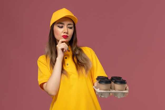 Front view of female courier in yellow uniform and cap holding brown delivery coffee cups thinking on pink wall