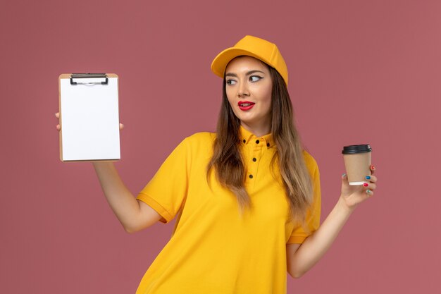 ピンクの壁に配達コーヒーカップとメモ帳を保持している黄色の制服とキャップの女性の宅配便の正面図