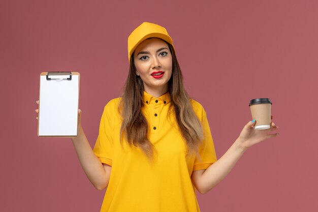 Вид спереди курьера-женщины в желтой униформе и кепке, держащего чашку кофе и блокнот на розовой стене