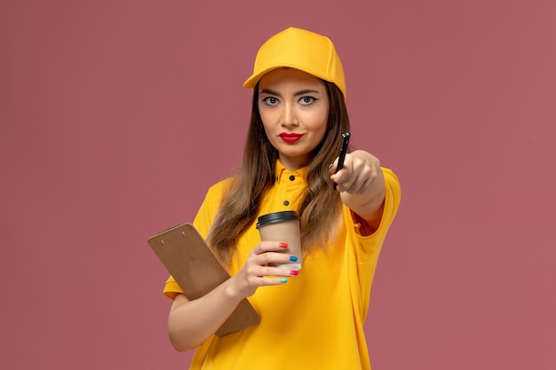 Front view of female courier in yellow uniform and cap holding delivery coffee cup and notepad with pen on pink wall