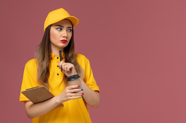 Front view of female courier in yellow uniform and cap holding delivery coffee cup and notepad with pen on pink wall