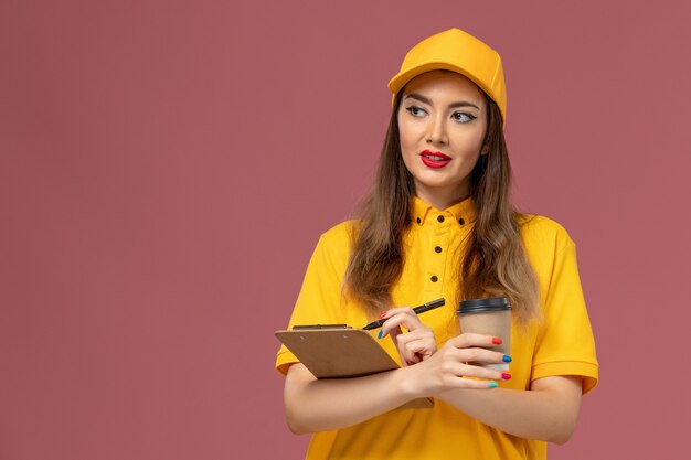 Front view of female courier in yellow uniform and cap holding delivery coffee cup and notepad with pen on pink wall