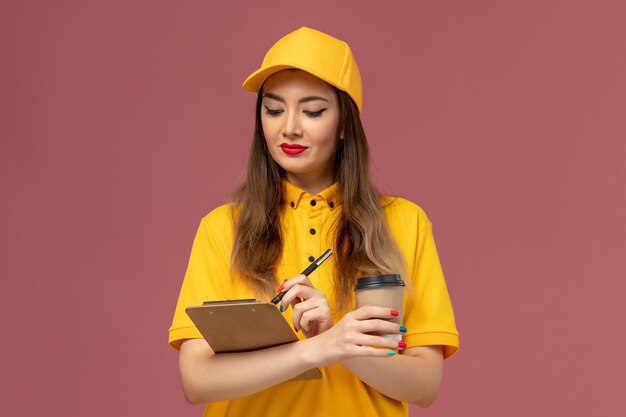 Front view of female courier in yellow uniform and cap holding delivery coffee cup and notepad with pen on pink wall