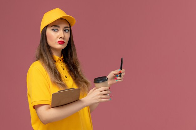 분홍색 벽에 펜으로 노란색 유니폼과 모자 배달 커피 컵과 메모장을 들고 여성 택배의 전면보기