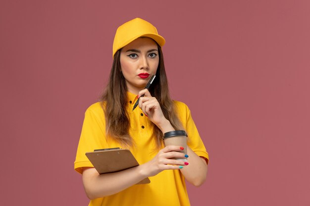 Front view of female courier in yellow uniform and cap holding delivery coffee cup and notepad with pen on the pink wall