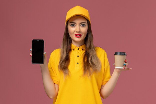 Front view of female courier in yellow uniform and cap holding delivery coffee cup and phone on pink wall