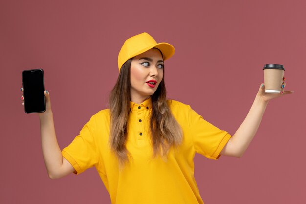 Front view of female courier in yellow uniform and cap holding delivery coffee cup and phone on pink wall